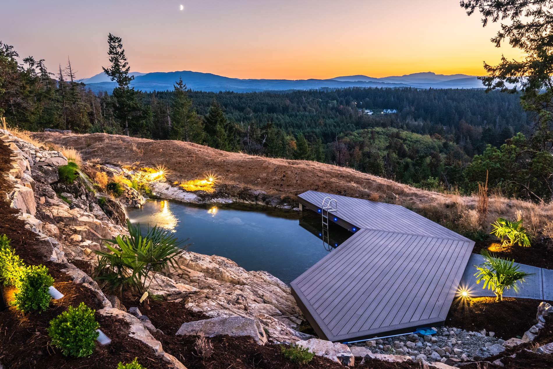 Natural swimming pond lit at dusk with a timber boardwalk and rock edging